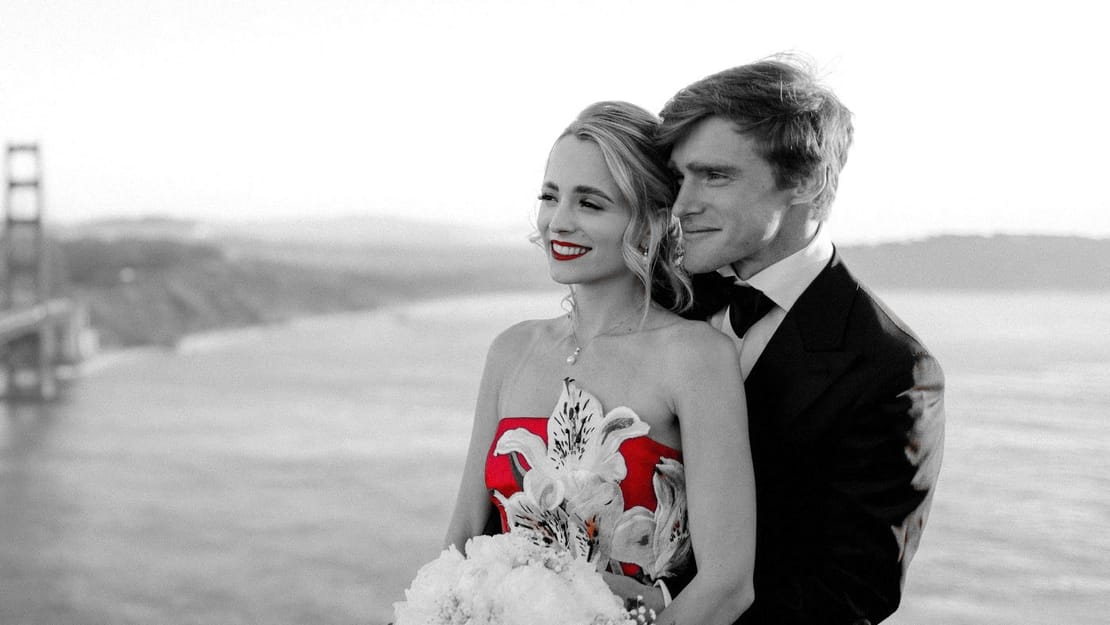 Maria on a wooden dock at sunset in Bora Bora, wearing a red gown and a flower crown, holding out her hand to show her engagement ring. Overwater bungalows sit in the background. The photo is black and white except for the red.
