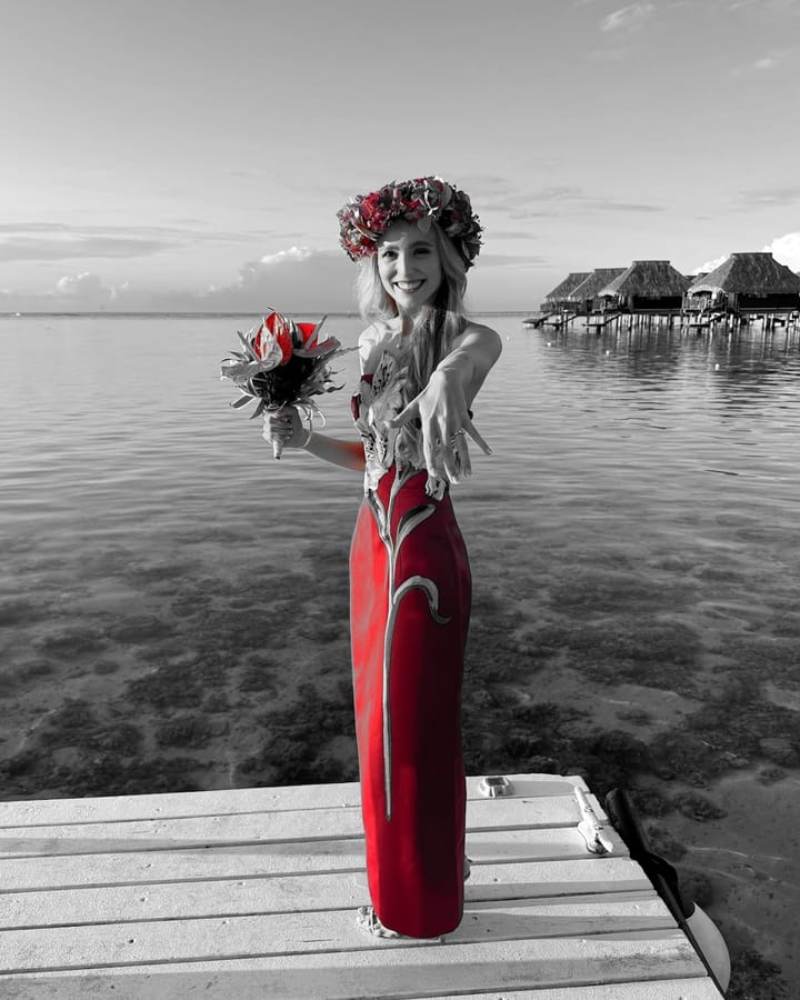 Maria on a wooden dock at sunset in Bora Bora, wearing a red gown and a flower crown, holding out her hand to show her engagement ring. Overwater bungalows sit in the background. The photo is black and white except for the red.